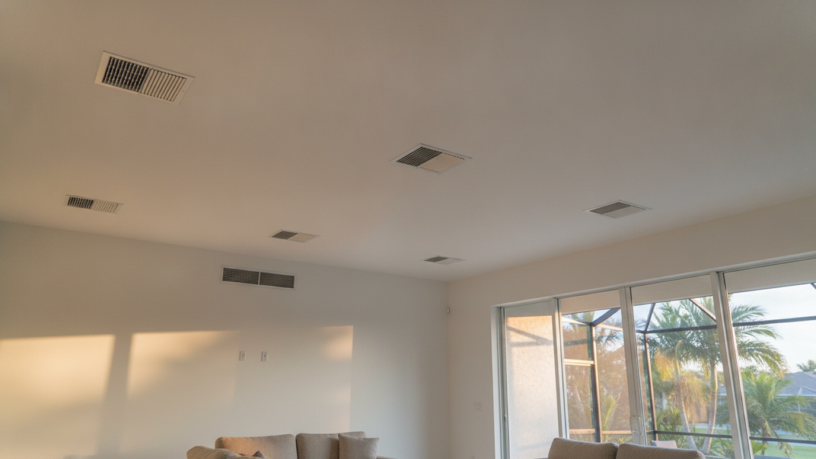 Several white ceiling supply registers and a wall return grille in the open living area of a single-story Englewood home, with a screened lanai and palms through the sliding glass doors